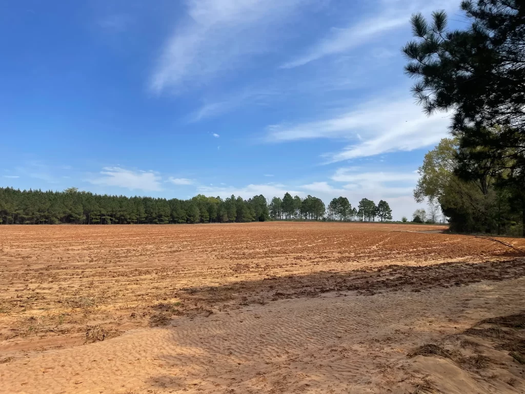 A large plowed field stretching into the distance, with a forested area in the background under a partly cloudy sky.
